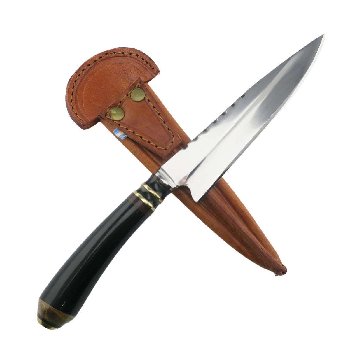 A Cow 6.3in Dark stainless steel knife with a black handle and brass accents, featuring a sharp, pointed blade with serrations on the spine, partially sheathed in a tan leather holster with brass rivets.